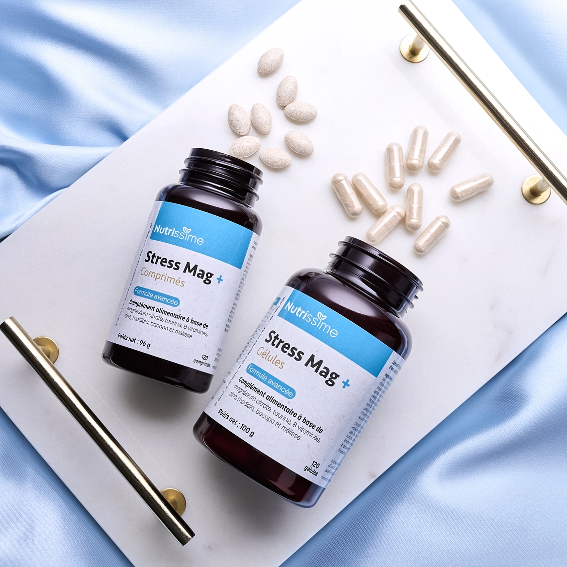 Photographie lifestyle de deux flacons de compléments alimentaires, contre le stress, couchés sur un plateau en marbre blanc, posé sur un lit avec un tissu en satin bleu ciel, avec des comprimes devant les flacons - Photographe lifestyle cosmétiques - photographe compléments alimentaires - photographe produits cosmétiques - Aurore Aivrest