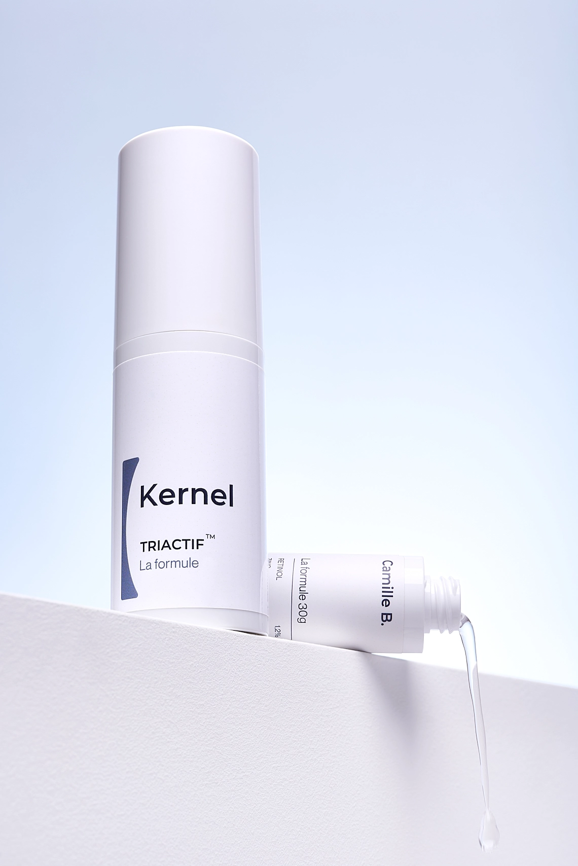 Photographie publicitaire d'un flacon de serum et sa recharge ouverte en train de se deverser. Le flacon et la recharge sont blancs avec des écritures bleu fonce. Ils sont poses sur un bloc blanc, le flacon est debout, a gauche et la recharge est couchee a l'arriere droite. Le fond est bleu ciel avec un halo lumineux, donnant un rendu assez clinique - Photographe cosmetiques - photographe produits cosmetiques - photographe publicitaire cosmetiques - Aurore Aivrest