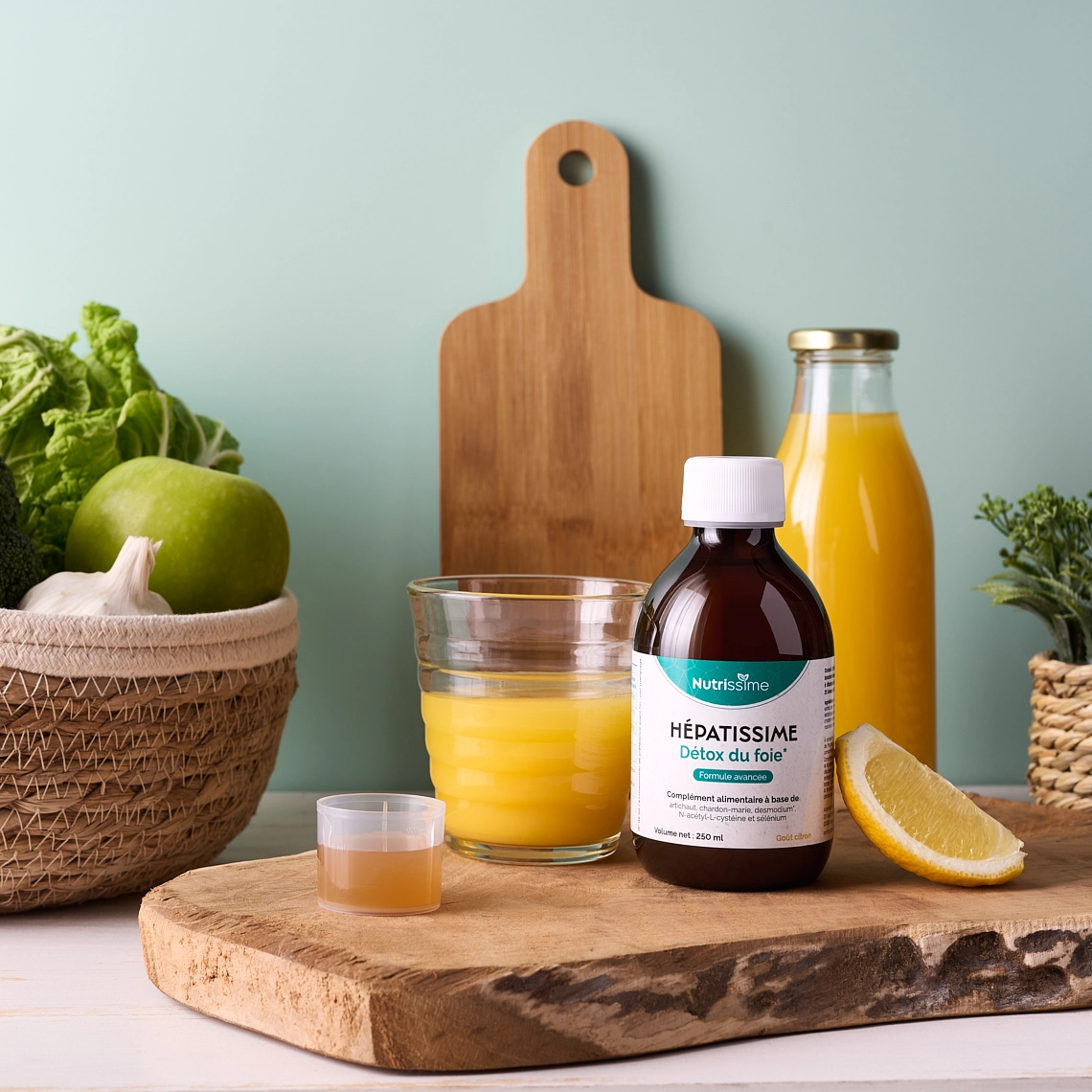 Photographie lifestyle d'une bouteille d'un complément alimentaire detox du foie sous forme liquide, sur une planche de bois, posee sur un plan de travail en bois peint en blanc, avec un verre de jus d'orange à côté du flacon, ainsi qu'un quartier de citron et le bouchon doseur rempli du produit. En arrière-plan il y a une plante verte dans un pot tressé, une bouteille en verre contenant du jus d'orange, une planche à decouper en bois debout adossée au mur, et une corbeille de fruits tressee contenant des fruits et légumes verts et une gousse d'ail. Le mur du fond est vert pastel. Le decor represente une cuisine - Photographe lifestyle cosmétiques - photographe compléments alimentaires - photographe produits cosmétiques - Aurore Aivrest