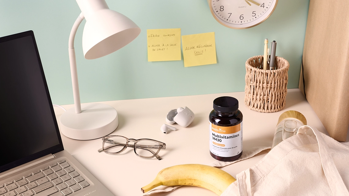 Photographie lifestyle d'un flacon de compléments alimentaires multivitamines posé sur un bureau beige. Le mur du fond est vert pastel. Le decor represente un bureau. Sur le bureau on voit un ordinateur portable, des lunettes de vue, une lampe de bureau, des ecouteurs sans fil, une pochette à documents, un pot à crayons. Il y a egalement un tote bag, avec son contenu qui depasse, notamment une banane et une gourde en verre contenant de l'eau. Sur le mur il y a une horloge blanche et doree ainsi que deux post-it indiquant des taches a faire apres la journee de travail - Photographe lifestyle cosmétiques - photographe compléments alimentaires - photographe produits cosmétiques - Aurore Aivrest
