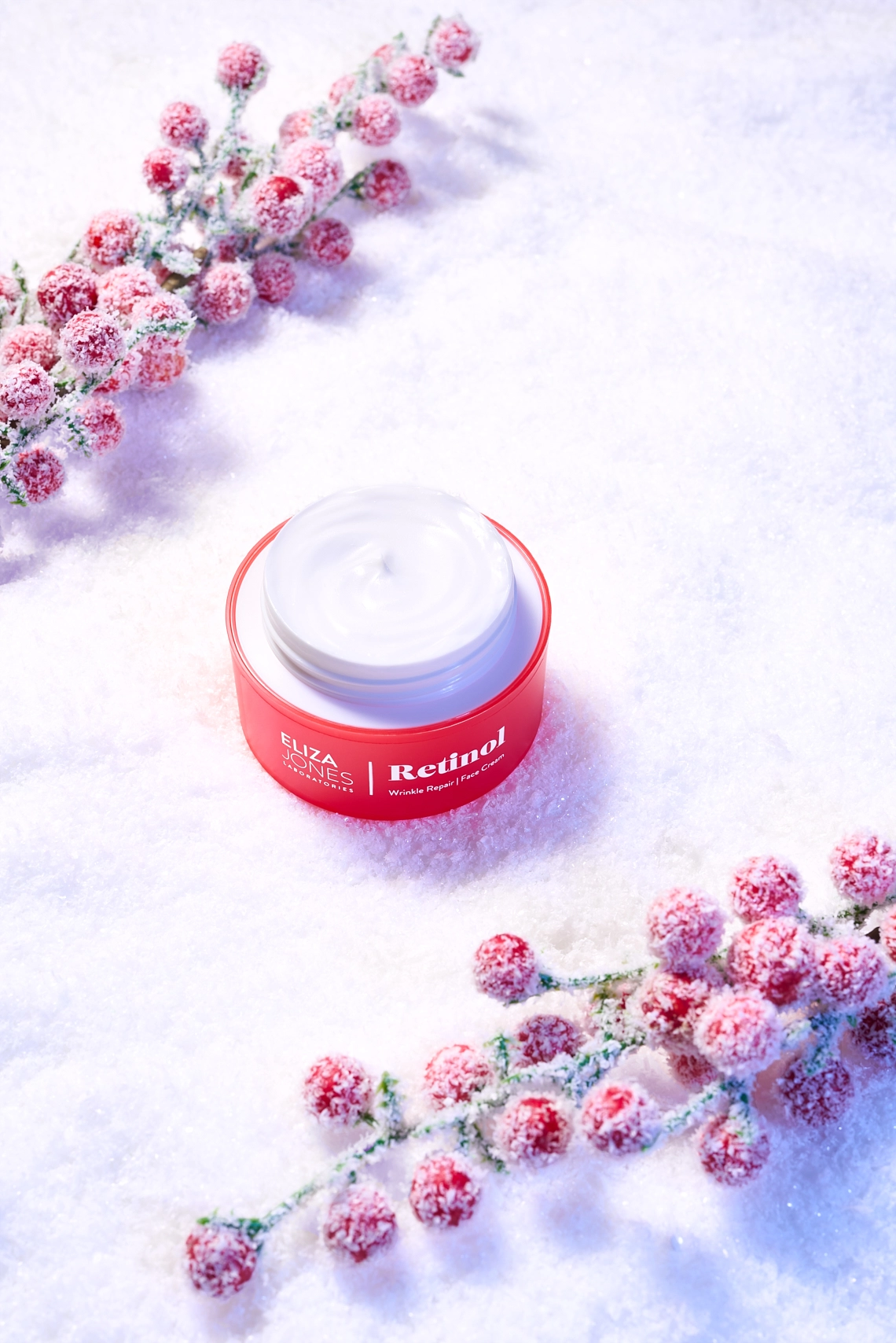 Photographie d'un pot de creme pour le visage pose dans la neige. Le pot de creme est ouvert et est de couleur rouge orange et s'accorde avec des branches a boules rouges enneigees. Theme hiver - Photographe cosmetiques - photographe publicitaire cosmetiques - photographe produits cosmetiques - Aurore Aivrest