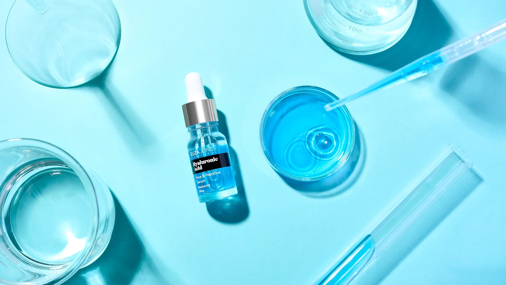 Photographie flatlay d'un serum acide hyaluronique pose sur un fond bleu. On y voit du materiel de laboratoire tels que becher, erlenmeyer, tube a essai, boite de petri, entonnoir. Ces accessoires creent des jeux d'ombres et de lumieres. Il y a egalement une pipette qui laisse couler du produit dans une boite de petri. Theme laboratoire - Photographe cosmetiques - photographe publicitaire cosmetiques - photographe produits cosmetiques - Aurore Aivrest