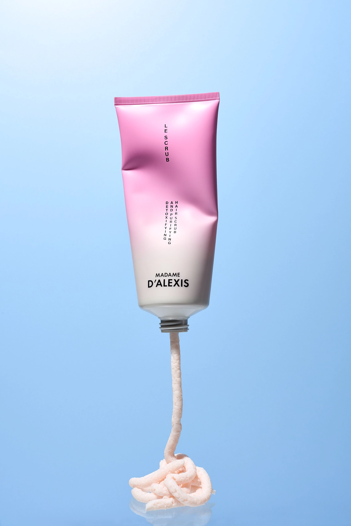 Photographie d'un tube de scrub gommage capillaire rose et blanc qui vole. Il est ecrase comme si une main invisible le pressait pour en extraire de la matiere. On voit justement la texture du produit qui sort et coule du tube. Le tout est sur un fond bleau ciel coherent avec cette mise en scene en levitation. Le rendu est moderne, dynamique est tendance en ce moment - Photographe cosmetiques - photographe publicitaire cosmetiques - photographe produits cosmetiques - Aurore Aivrest
