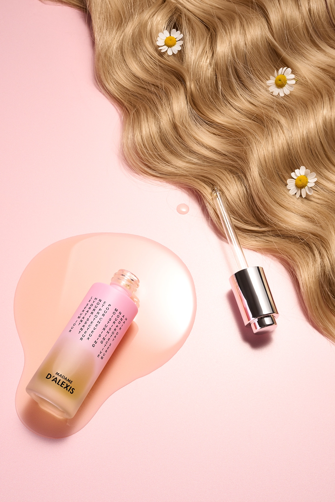 Photographie d'un serum de jour pour les cheveux pose sur un fond rose. On y voit le serum se deverser sur le fond creant une flaque. On voit egalement des cheveux blonds, dans lesquels se trouvent des fleurs de camomille matricaire. Ainsi que le bout de la pipette du serum. - Photographe cosmetiques - photographe publicitaire cosmetiques - photographe produits cosmetiques - Aurore Aivrest