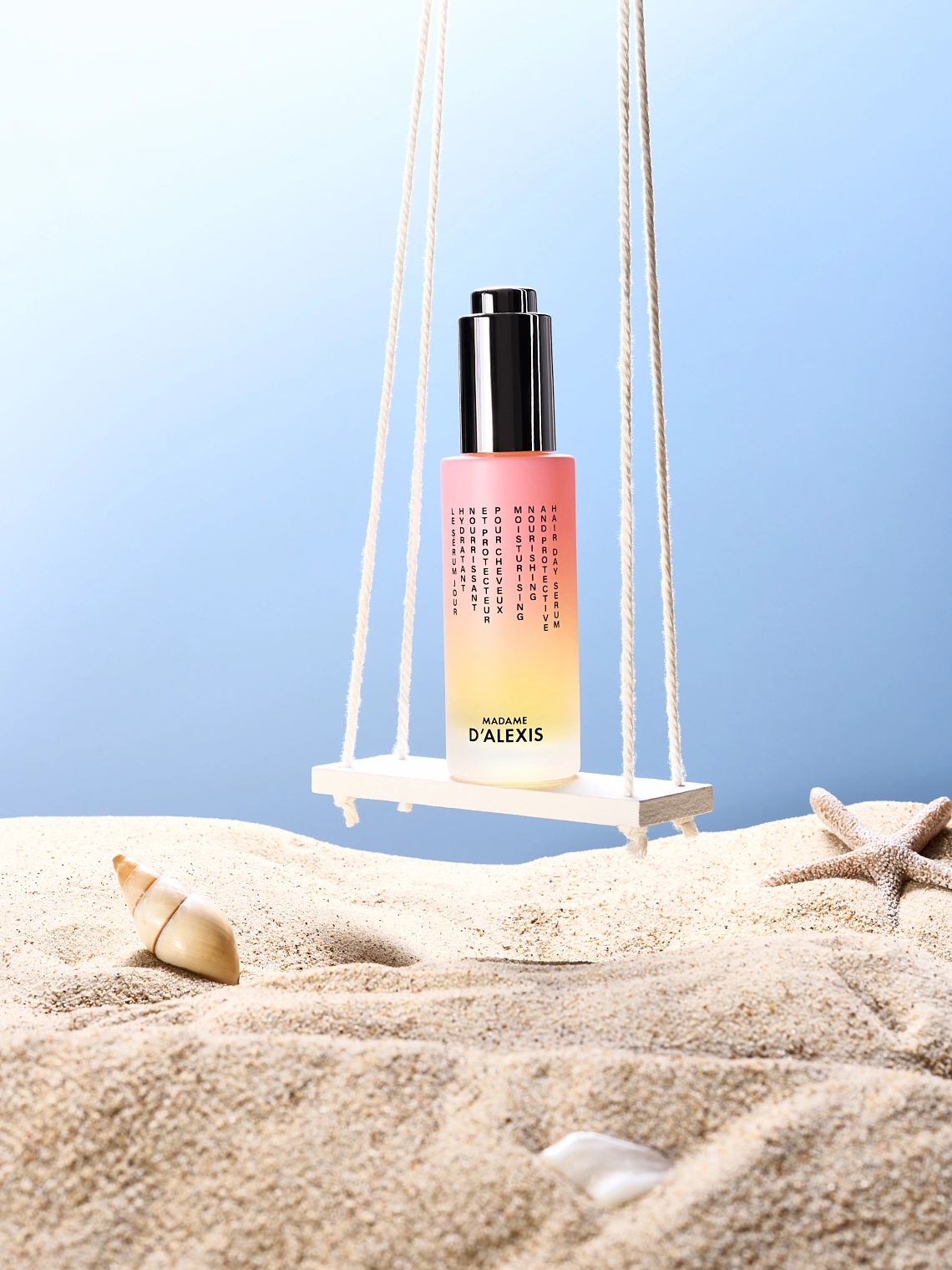 Photographie d'un flacon de serum de jour pour les cheveux. Le flacon est rose et jaune. Le flacon est debout sur une balancoire sur la plage. On y voit un ciel bleu et du sable avec des coquillages et etoile de mer. Ambiance ete, summer, plage, vacances - Photographe cosmetiques - photographe publicitaire cosmetiques - photographe produits cosmetiques - Aurore Aivrest