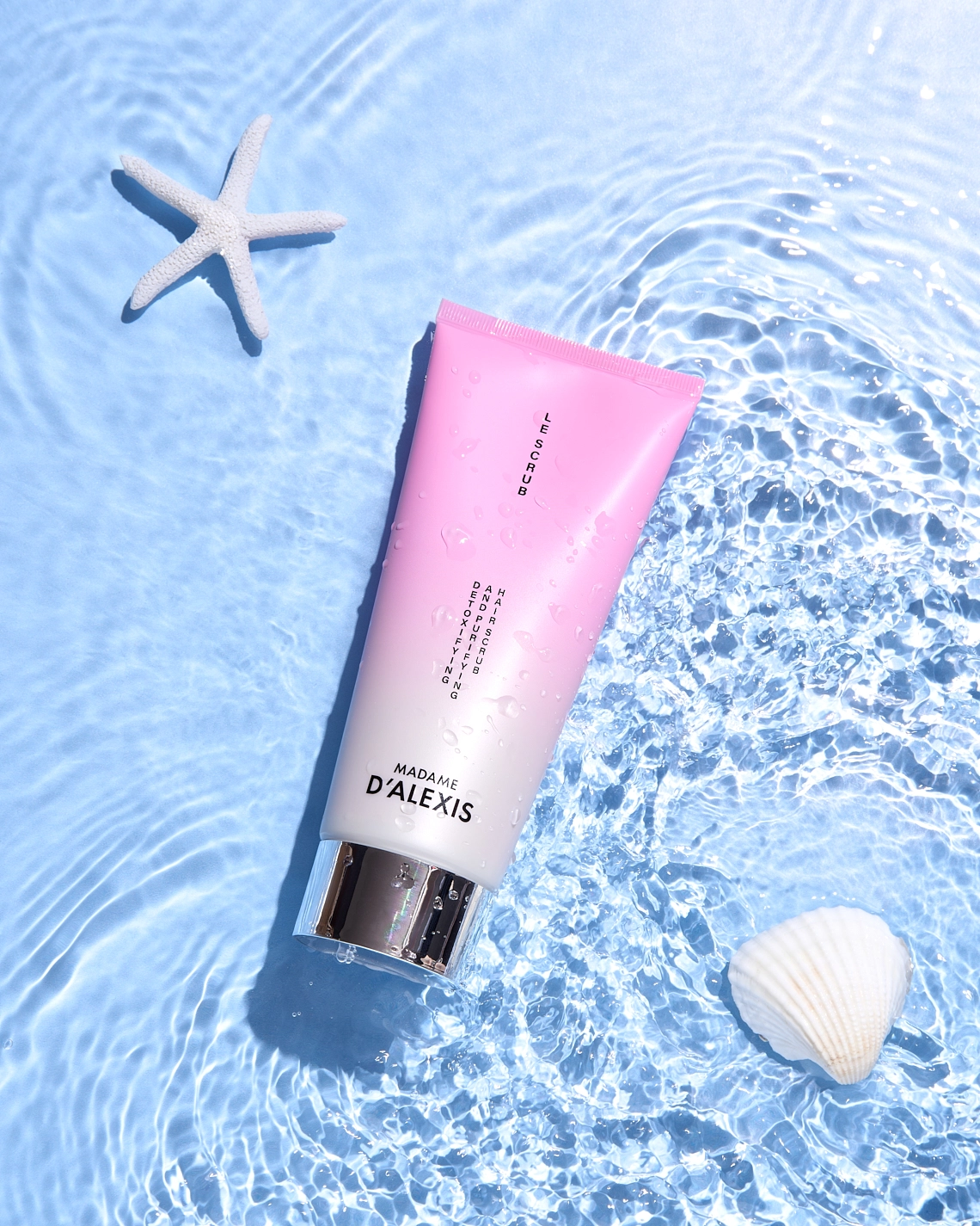 Photographie d'un tube de scrub gommage capillaire rose et blanc couche dans l'eau. On y voit egalement une etoile de mer blanche et un coquillage blanc, pour rappeler que c'est de l'eau de mer. Le fond est bleu pour qu'on comprenne bien qu'il s'agisse d'eau. Le rendu est luxueux, délicat, frais et mouvemente. - Photographe cosmetiques - photographe publicitaire cosmetiques - photographe produits cosmetiques - Aurore Aivrest