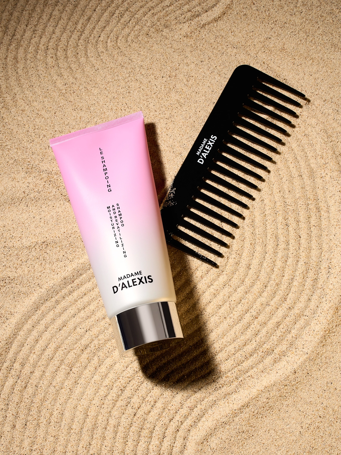 Photographie d'un tube de shampooing rose et blanc avec un peigne noir, couches sur du sable. Il y a des traces en zig zag dans le sable, creees avec le peigne. Elles apportent un rendu graphique, rappellent les meches de cheveux puisqu'il s'agit de produits capillaires, et rappellent les jardins zen japonais. Le rendu est graphique et contraste - Photographe cosmetiques - studio photo cosmetiques - photographe publicitaire cosmetiques - Aurore Aivrest