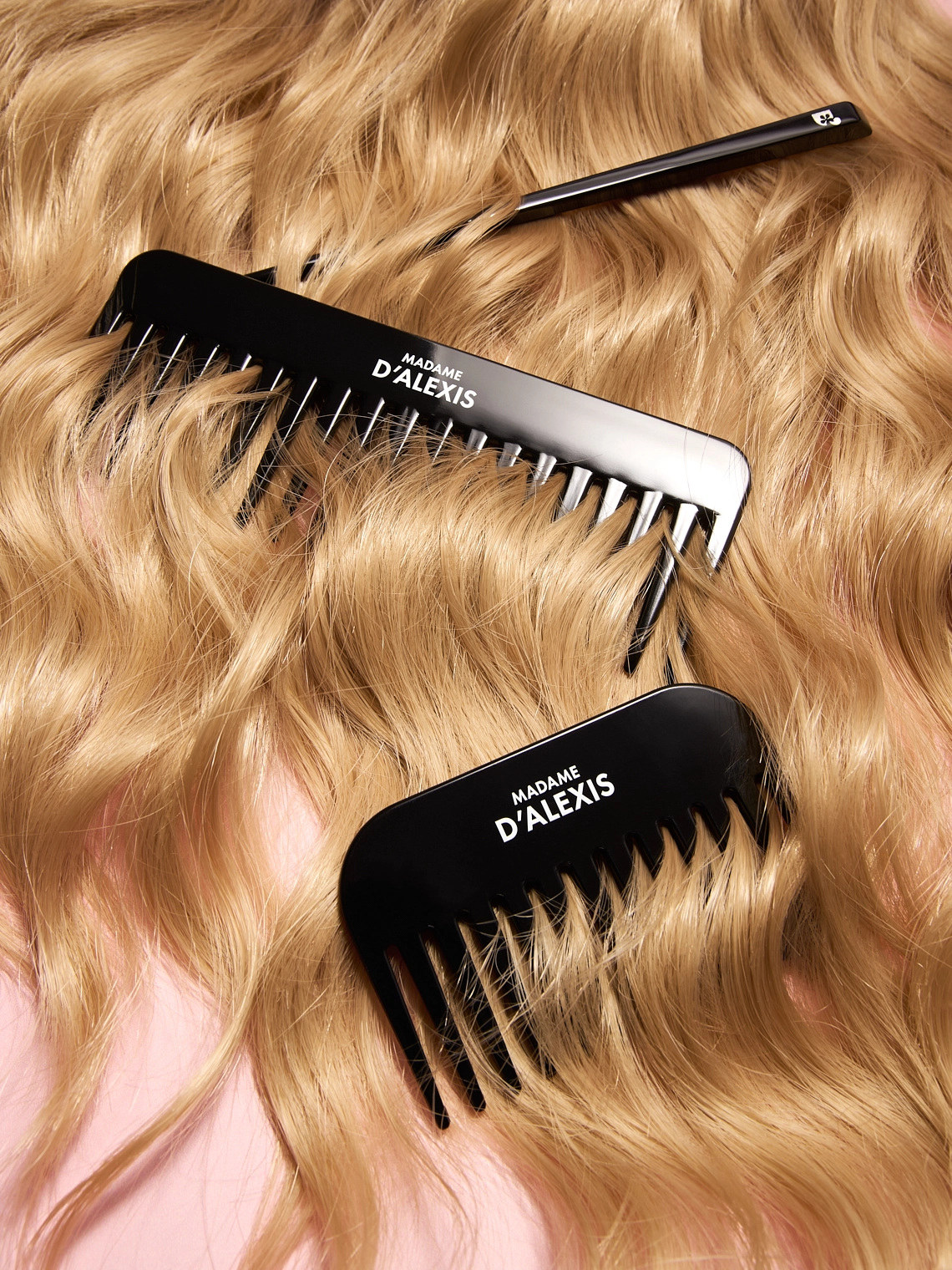 Photographie de trois accessoires capillaires. Il s'agit d'un peigne court, d'un peigne long et d'un pic a cheveux. Tous sont de couleur noire, avec le logo de la marque en blanc. Ils sont disposes dans des cheveux blonds ondules. Le tout sur un fond rose clair. Ambiance coiffeur, capillaire, moderne, grand angle. Le rendu est assez luxueux - Photographe cosmetiques - photographe publicitaire cosmetiques - photographe produits cosmetiques - Aurore Aivrest