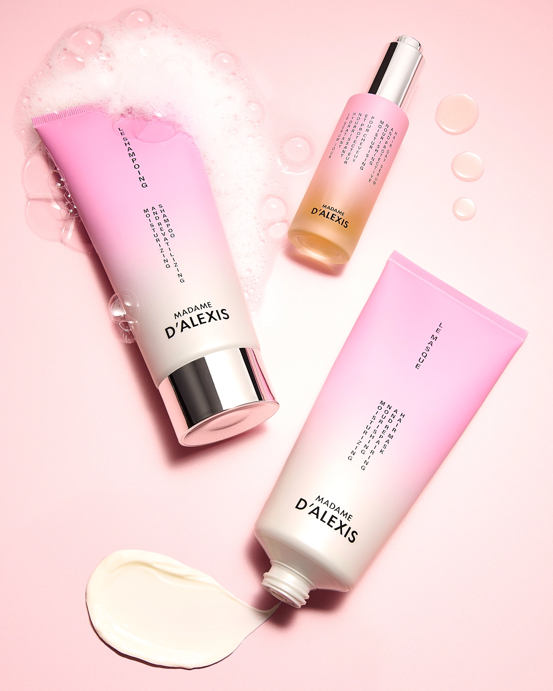 Photographie de trois produits capillaires : un tube de shampoing, un tube de masque et un flacon de serum jour. Les tubes dont rose et blanc, tandis que le flacon de serum est rose et jaune orange. Ils sont couches sur un fond rose clair. A cote de chacun de ces produits on y voit leur texture, comme de la mousse avec des bulles du shampoing, des gouttes de serum, un etalement du masque. Ambiance moderne, coloree, texturee, sensorielle. Le rendu est assez luxueux - Photographe cosmetiques - photographe publicitaire cosmetiques - photographe produits cosmetiques - Aurore Aivrest