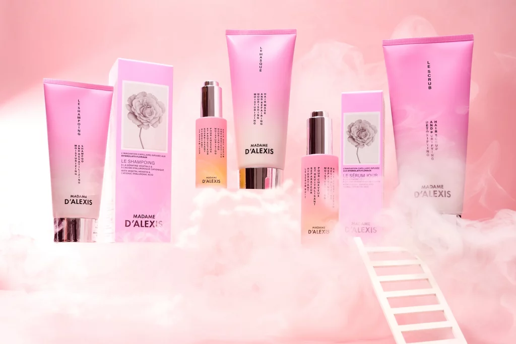 Photographie d'une gamme de produits capillaires : un tube de shampoing et sa boite, un tube de masque, un tube de scrub, un flacon de serum jour et sa boîte et un flacon de serum nuit. Les tubes, les boites et le flacon de serum nuit sont rose et blanc, tandis que le flacon de serum jour est rose et jaune orange. Ils sont debout sur les nuages. Une echelle blanche est positionnee pour monter sur ces nuages. Ambiance originale, moderne, coloree, inspirationnelle, delicate. Le rendu est assez luxueux - Photographe cosmetiques - photographe publicitaire cosmetiques - photographe produits cosmetiques - Aurore Aivrest